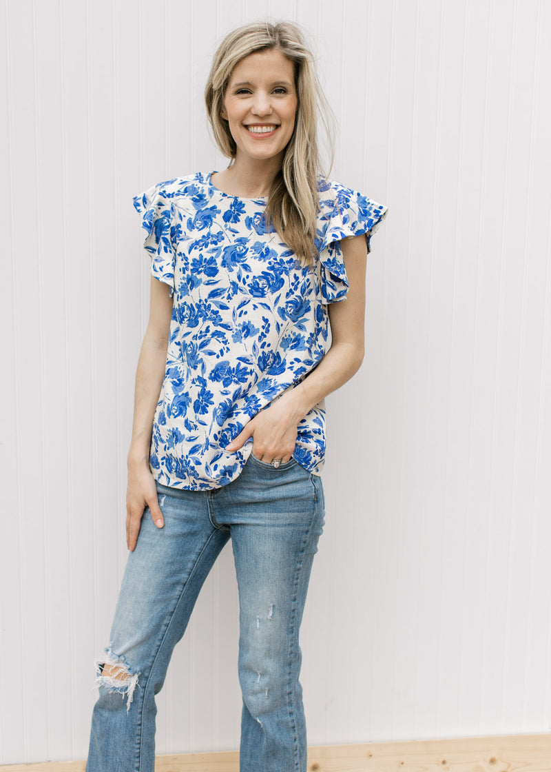 Model wearing jeans with a cream top with bright blue floral pattern and ruffle cap sleeves.