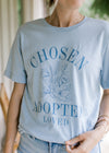 Model wearing a pale blue tee shirt with floral graphic and words chosen, adopted and loved. 