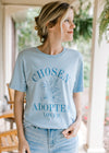 Model wearing jeans with a pale blue tee with floral graphic and words chosen, adopted and loved. 