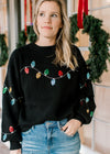 Model wearing a black sweater with colored sequins lights, long sleeves and a round neck. 