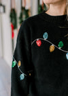 Detail view of colored sequins lights on a long sleeve black sweater with a round neck. 