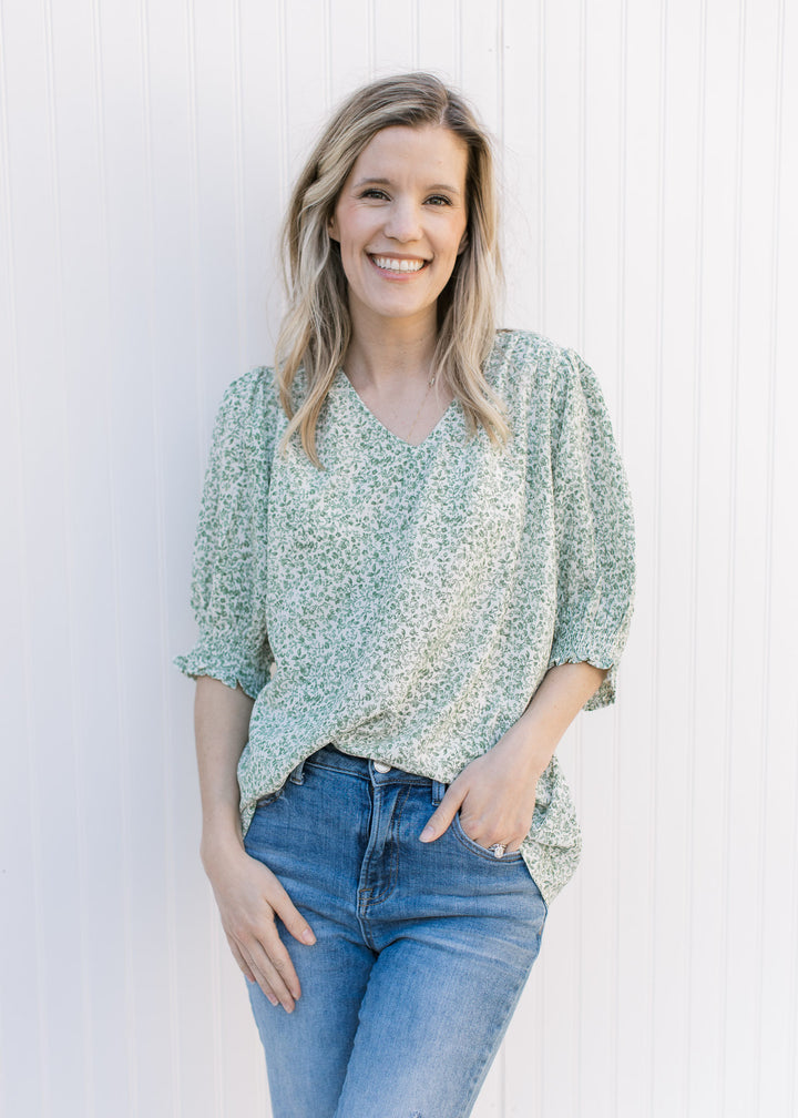 Model wearing jeans with a cream polyester top with green floral print and 3/4 bubble sleeves.
