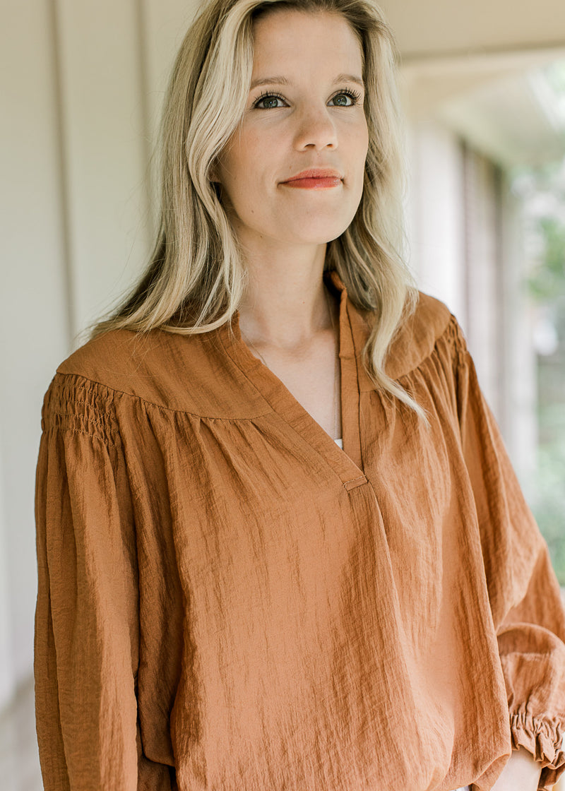 Model wearing a cinnamon blouse with smocked detail at shoulder long sleeves and v-neck. 