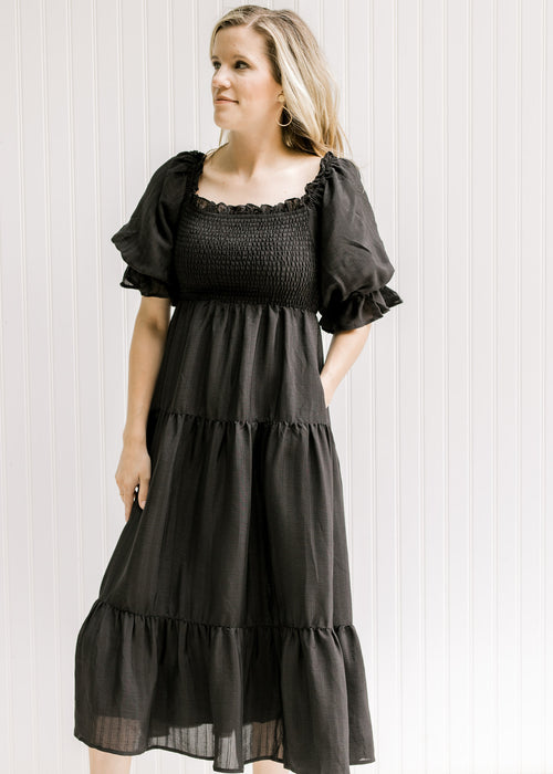 Model wearing a black tiered midi dress with a smocked bodice and short puff sleeves with pockets. 