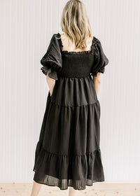 Back view of Model wearing a black tiered midi dress with a smocked bodice and short puff sleeves. 
