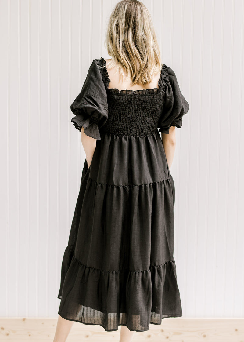 Back view of Model wearing a black tiered midi dress with a smocked bodice and short puff sleeves. 