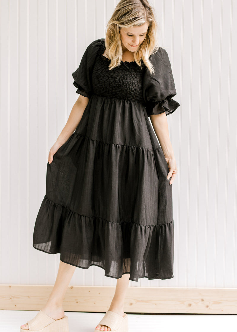 Model wearing wedges with a black tiered midi dress with a smocked bodice and short puff sleeves. 