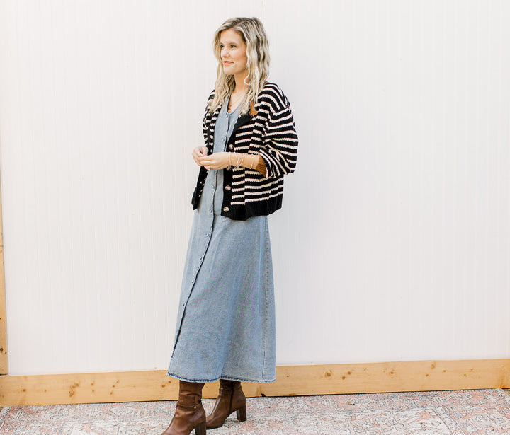 Woman wearing a striped cardigan, denim dress, and brown boots against a white wall.