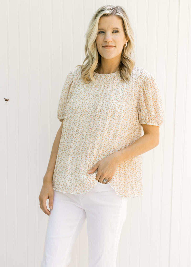 Model wearing white jeans with a pleated ivory top with a microfloral patter and short sleeves. 