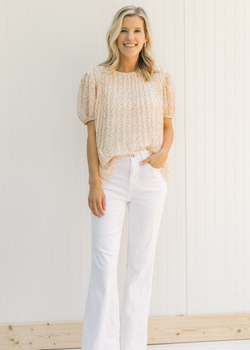 Model wearing white flare jeans with a pleated ivory microfloral top with short sleeves. 