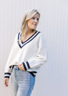 Model wearing jeans with a cream sweater with two navy stripes at neck, hem and cuff. 