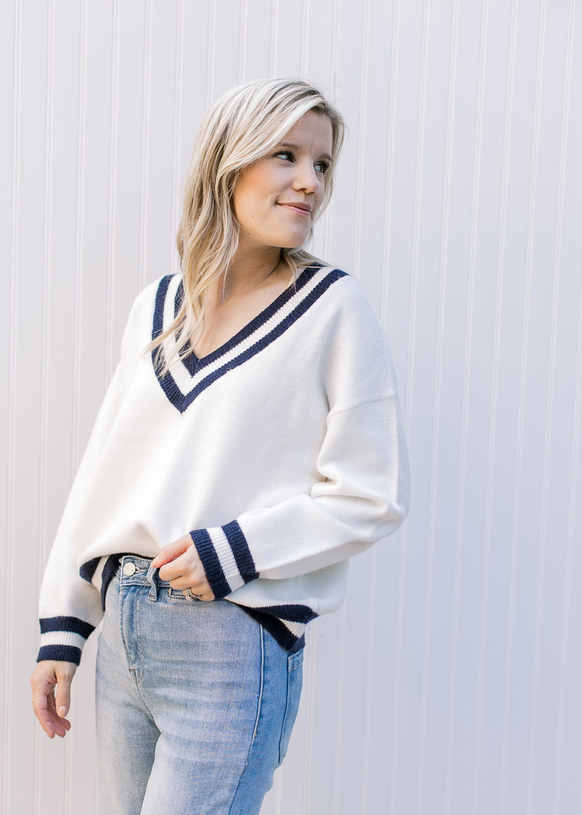 Model wearing jeans with a cream sweater with two navy stripes at neck, hem and cuff. 