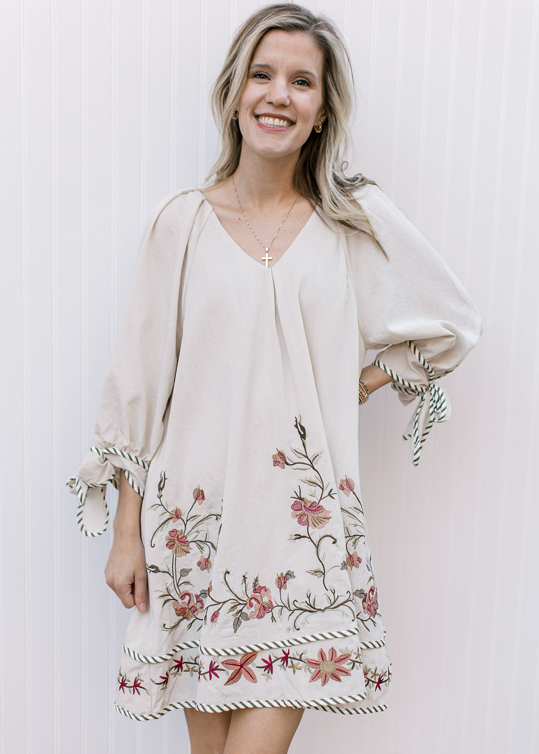 Model wearing a cream v-neck dress with green, gold and maroon embroidered flowers and 3/4 sleeves.