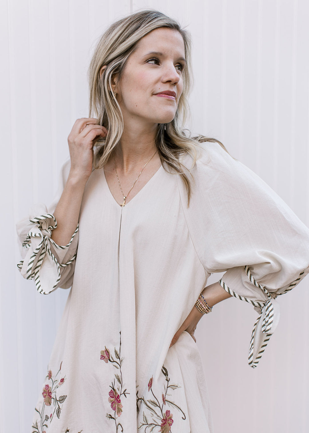 Model wearing a cream v-neck dress with 3/4 sleeves, pleating detail and floral detail. 