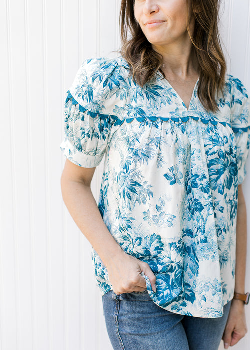 Model wearing a cream top with teal flowers, short sleeves and scallop detail.