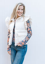 Model wearing a cream puffer vest with ruffle at shoulder, zipper and front patch pockets. 