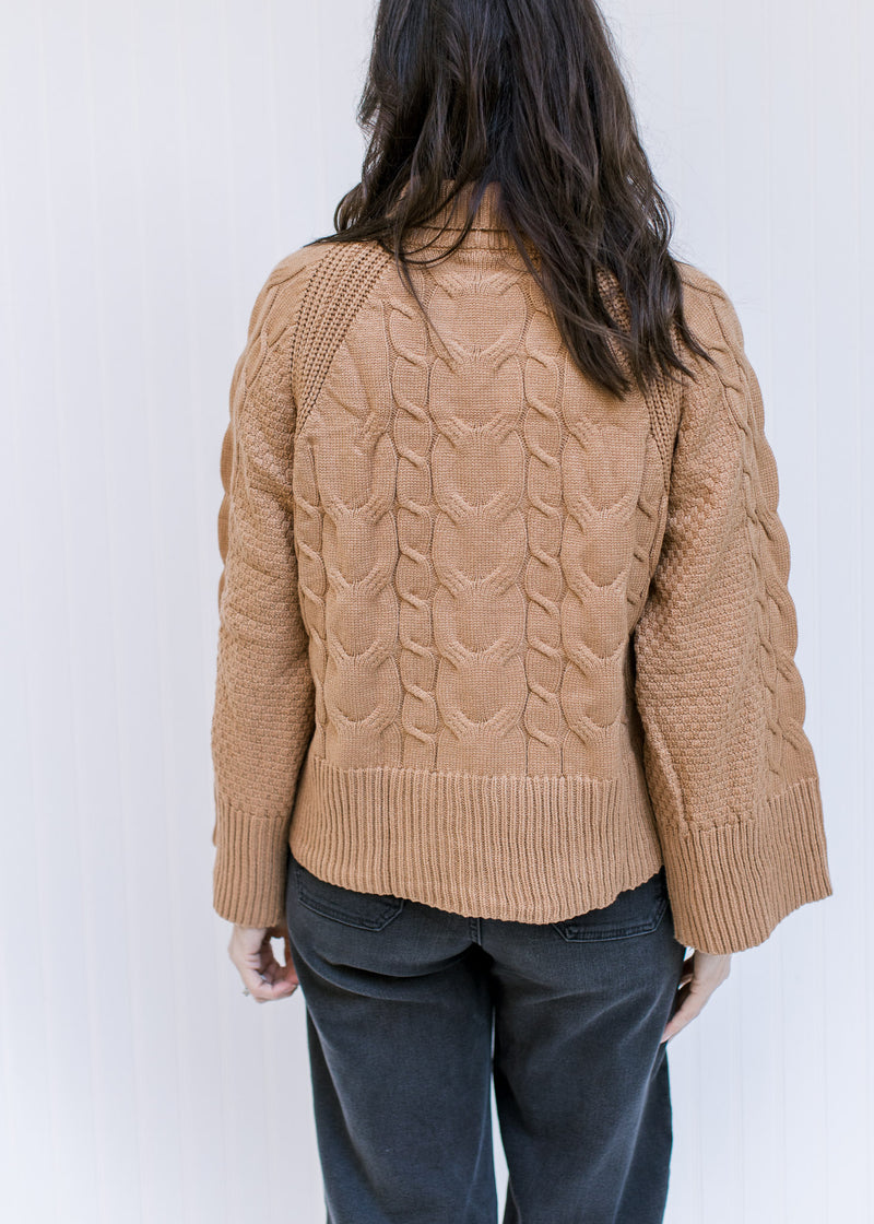 Back view of Model wearing a camel cable knit sweater with a turtle neck and long sleeves. 