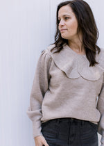 Model wearing a taupe sweater with an exaggerated collar and long sleeves with pleated shoulders. 
