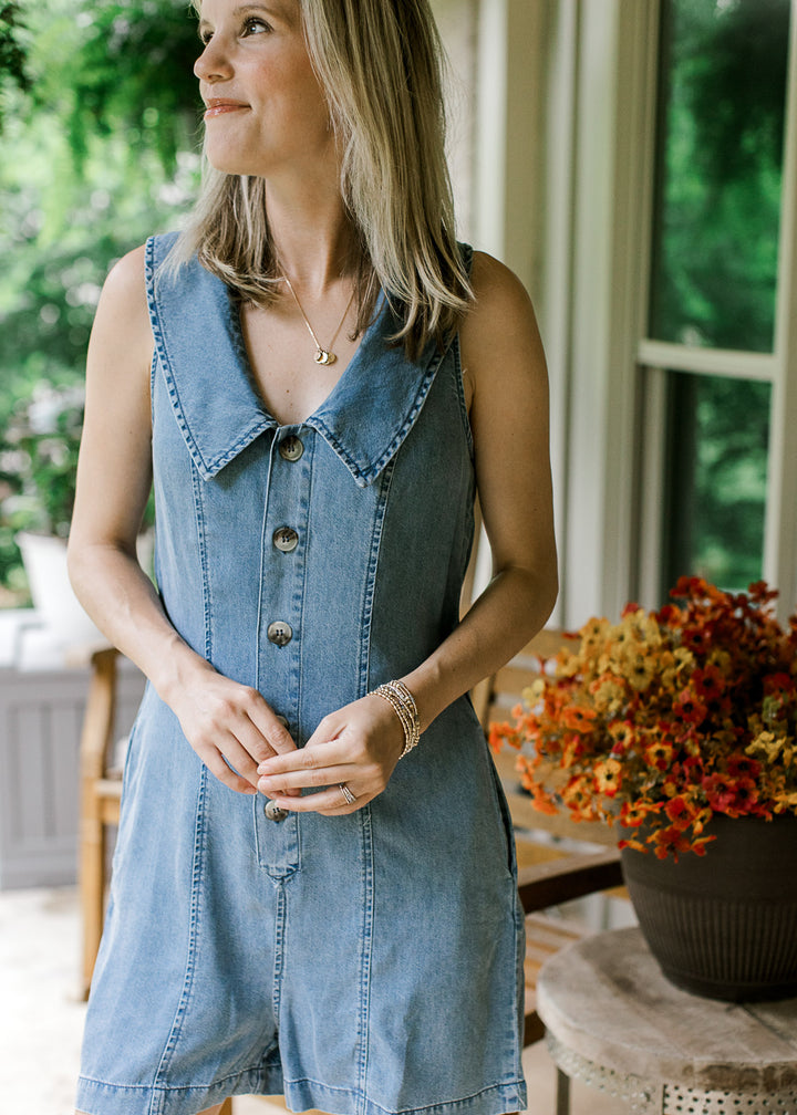 Close up of v-neck with flap collar on a sleeveless, medium wash denim romper with buttons. 
