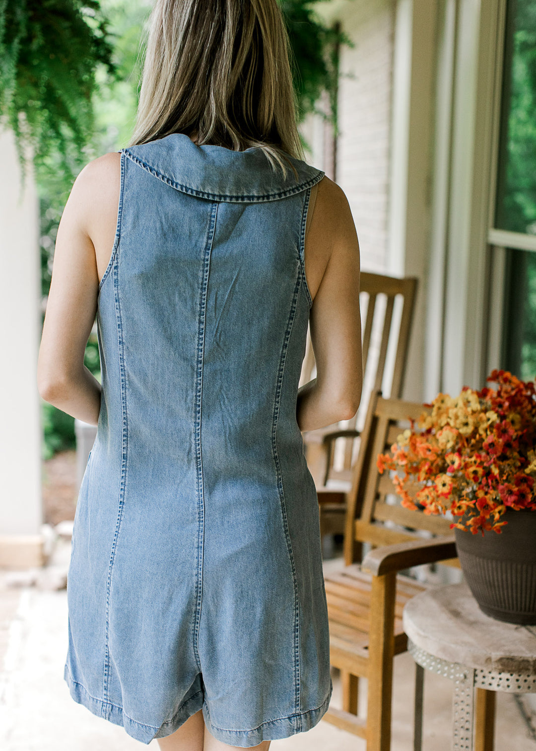Back view of Model wearing a sleeveless denim romper with collared v-neck and pockets. 