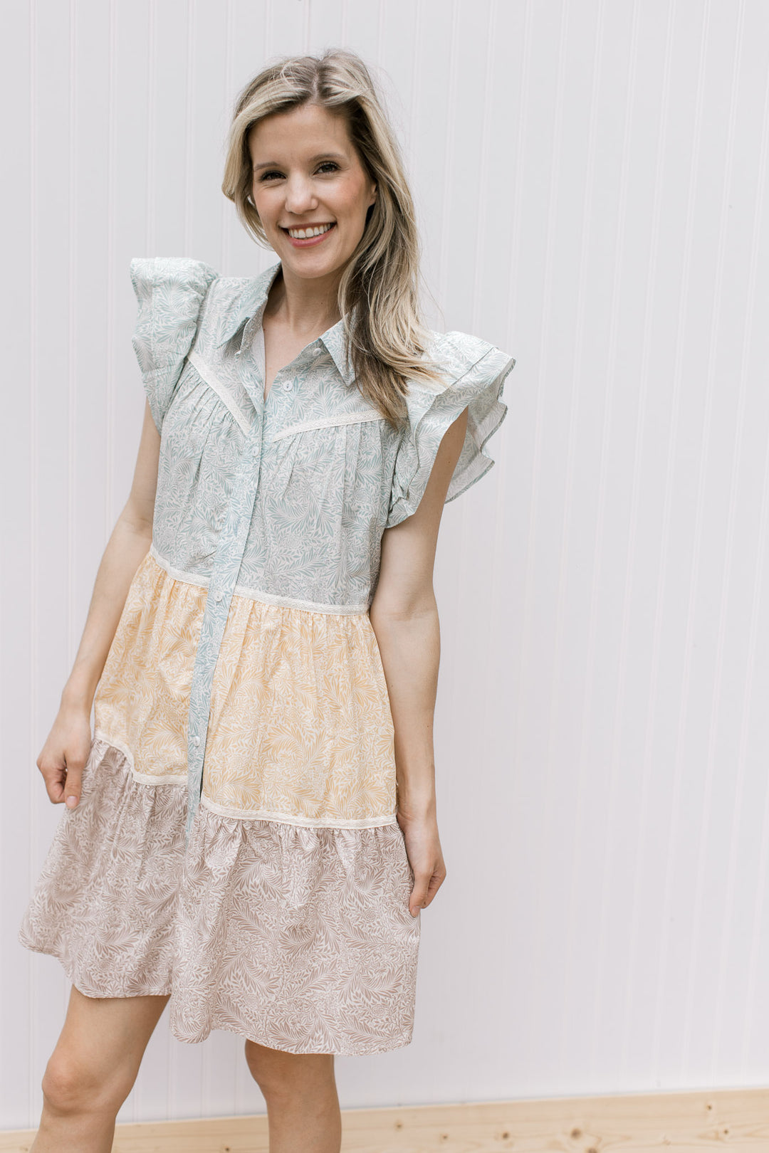 Model is wearing a sage, mustard and khaki floral dress with lace detail and ruffle cap sleeves. 