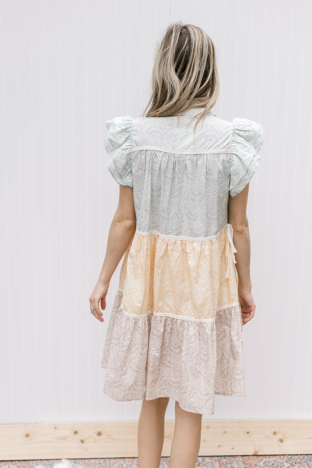 Back view of Model is wearing a sage, mustard and khaki floral dress with ruffle cap sleeves.