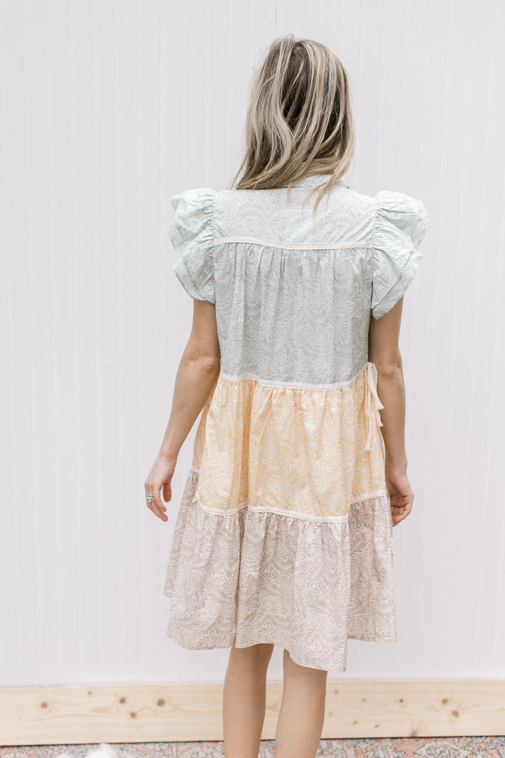 Back view of Model is wearing a sage, mustard and khaki floral dress with ruffle cap sleeves.