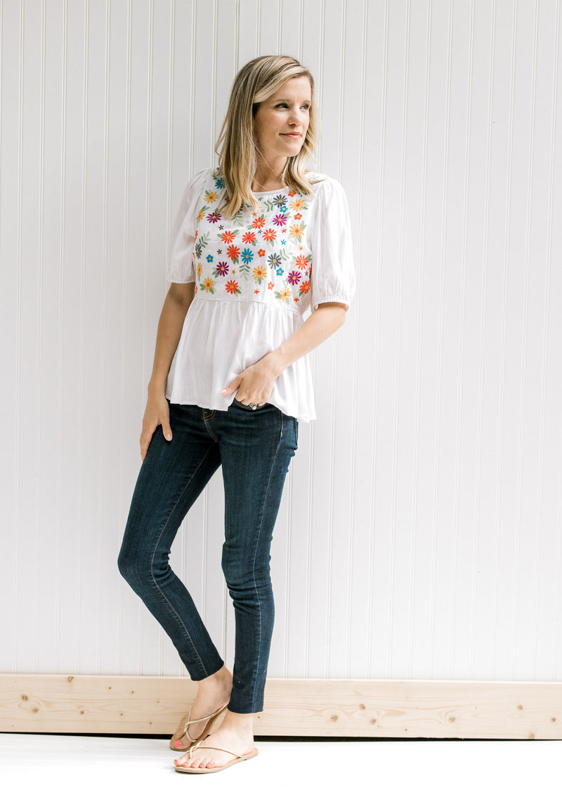 Model wearing jeans with an ivory babydoll top with embroidered floral pattern on bodice.