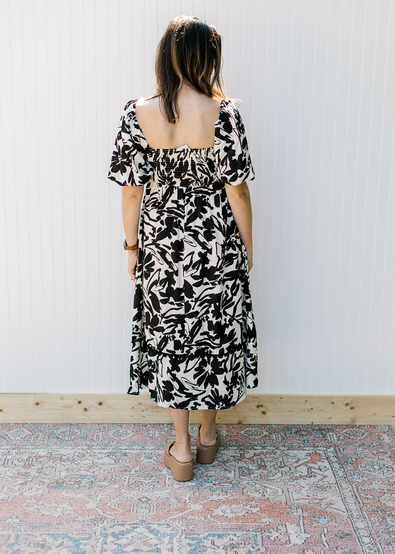 Back view of model wearing a cream midi with black flowers, square neck and short sleeves.