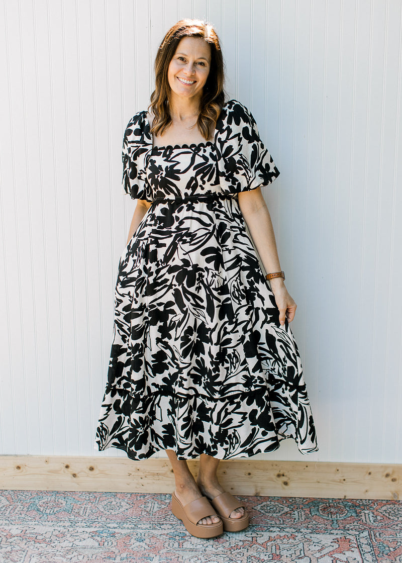 Model wearing wedges and a cream dress with black flowers and short sleeves.