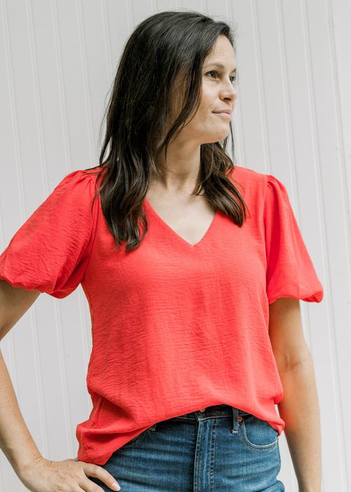 Model wearing a coral v-neck top with bubble short sleeves and pleated shoulder detail. 
