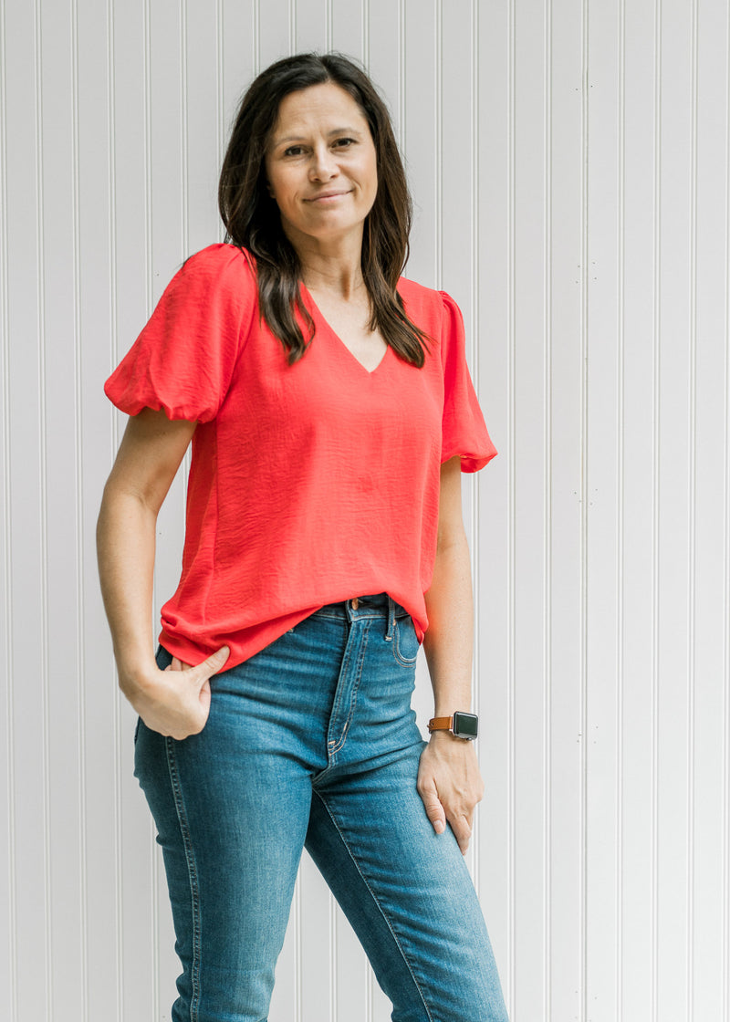Model wearing jeans with a coral v-neck top with bubble short sleeves and pleated shoulder detail. 