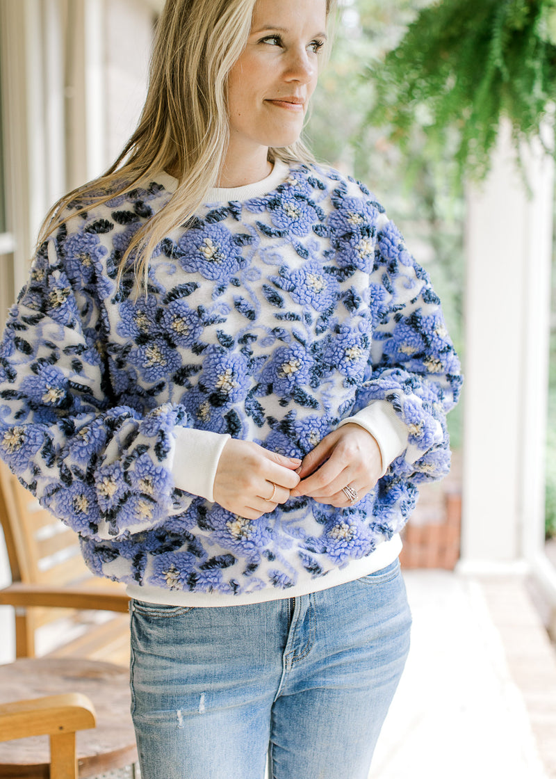 Model wearing a white sweater with bluish purple elevated flowers and long sleeves. 