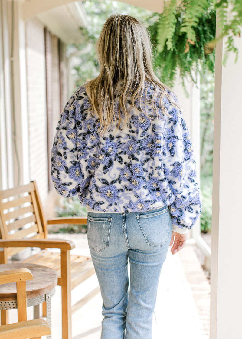 Back view of Model wearing a white sweater with bluish purple elevated flowers and long sleeves. 