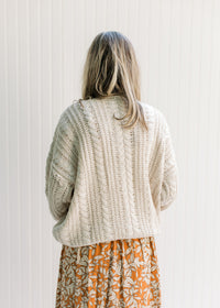 Back view of Model wearing a cream cable knit acrylic sweater with a v-neck and long sleeves.