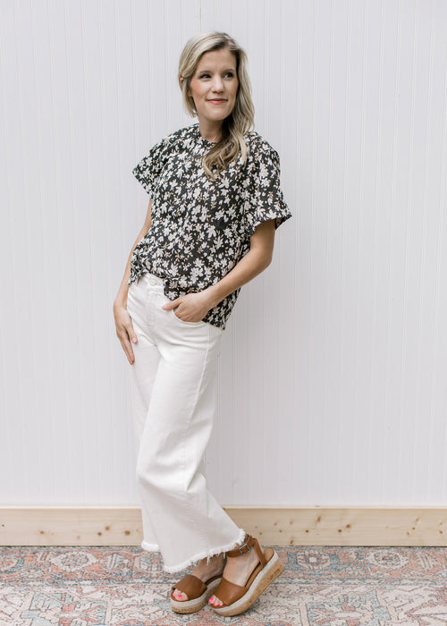 Model wearing white jeans, sandals and a black top with white daisies and short sleeves.