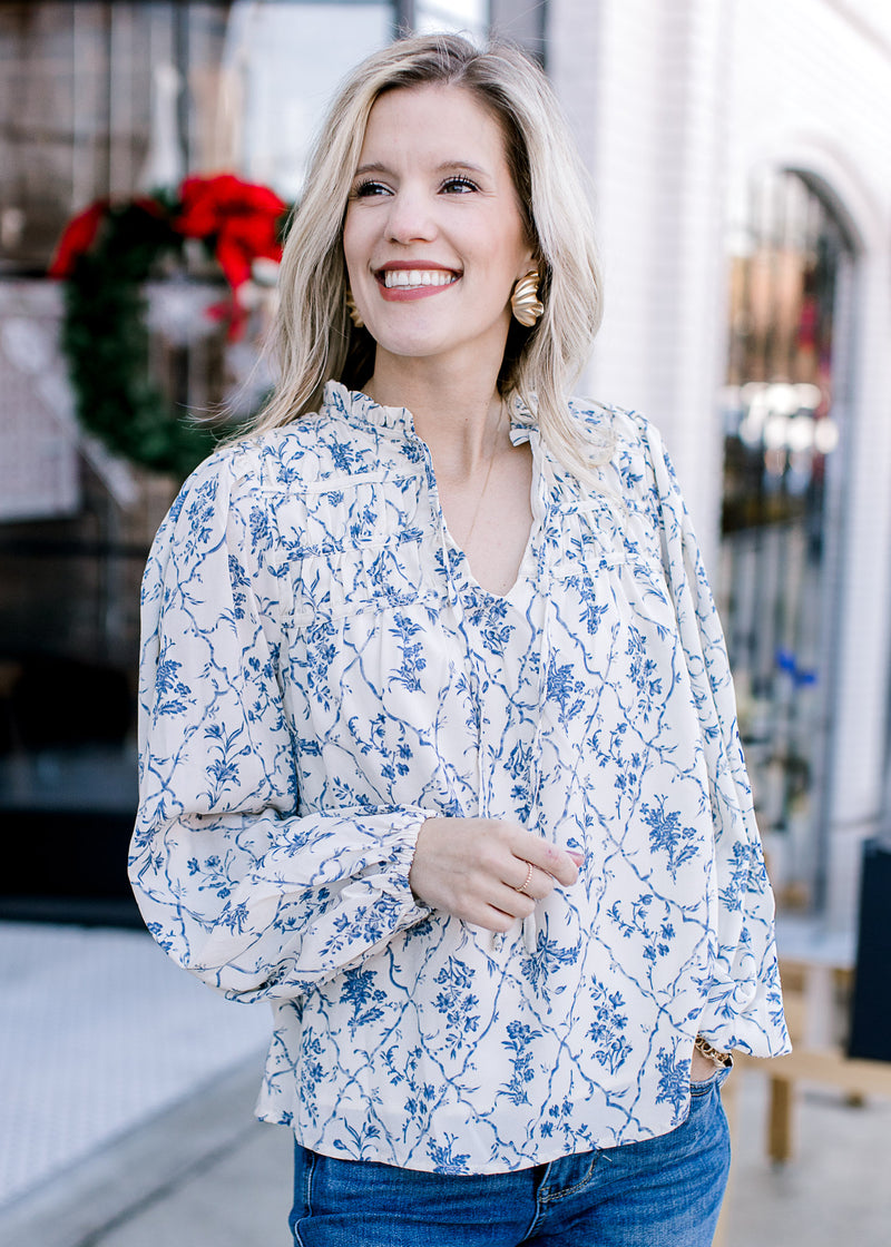 Model wearing a cream top with blue floral print, long bubble sleeve and ruffled neck with tie. 