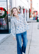 Model wearing jeans and a cream top with blue floral print, long sleeve and ruffled neck with tie. 