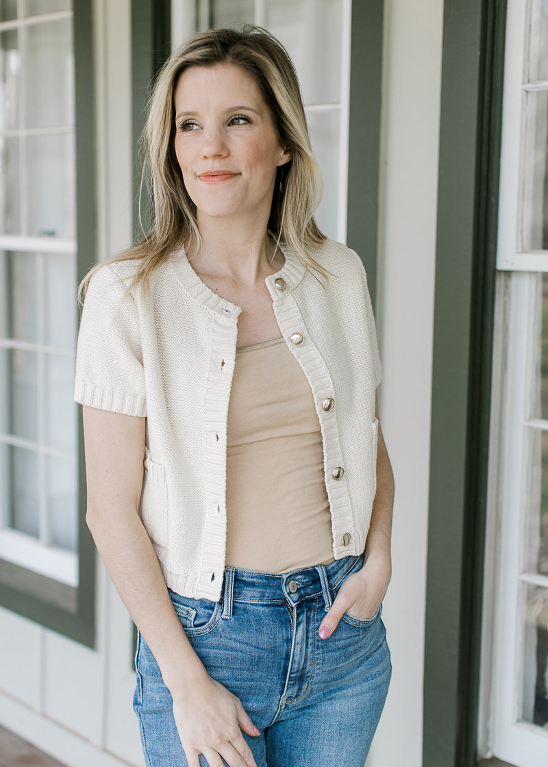 Model wearing a cream short sleeve sweater with gold buttons and patch pockets. 