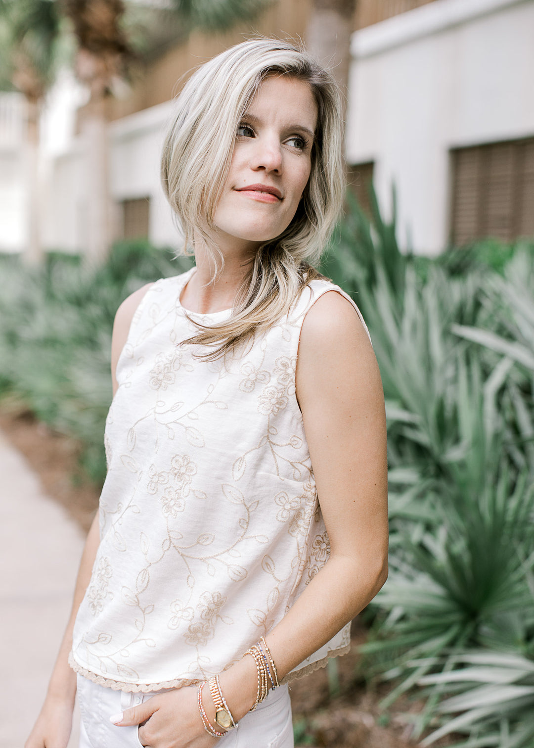 Model wearing an ivory sleeveless top with tan floral, round neck and tan scalloped trim.