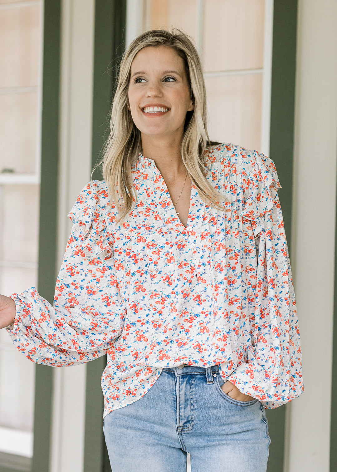 Model wearing a cream blouse with orange and blue floral, long sleeves, v-neck and ruffle shoulder.