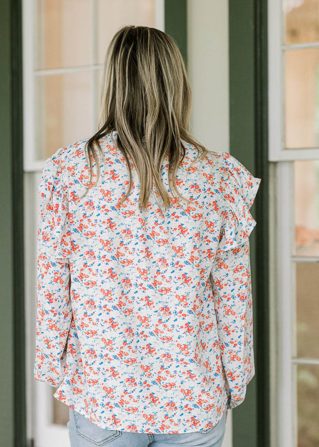 Back view of Model wearing a long sleeve cream top with orange and blue floral and ruffle shoulder.