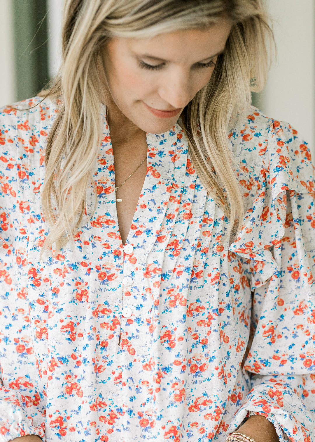 Model wearing an off white top with orange and blue floral, pleating and three button front. 