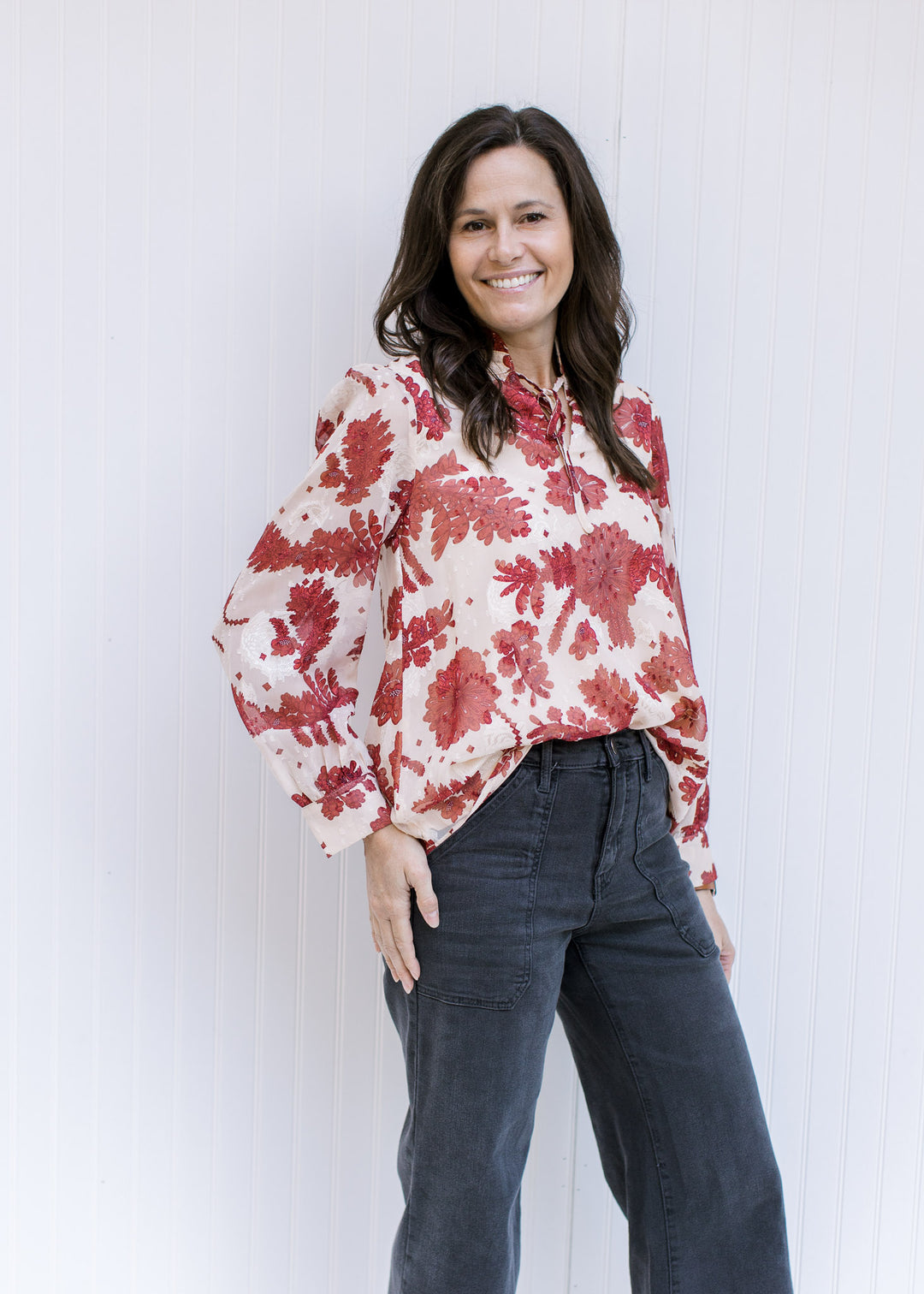 Model wearing a cream top with a rust floral pattern, sheer long sleeves and v-neck with a ruffle. 