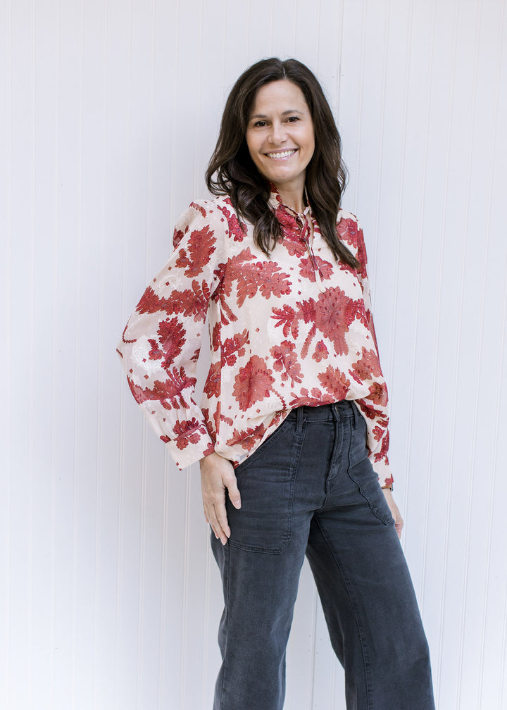 Model wearing a cream top with a rust floral pattern, sheer long sleeves and v-neck with a ruffle. 