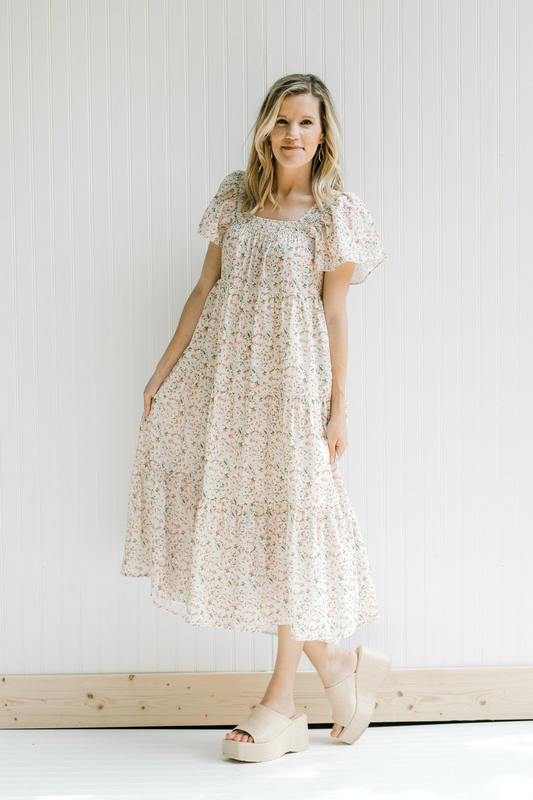 Blonde model wearing a tiered cream dress with a pink floral pattern and short flutter sleeves.