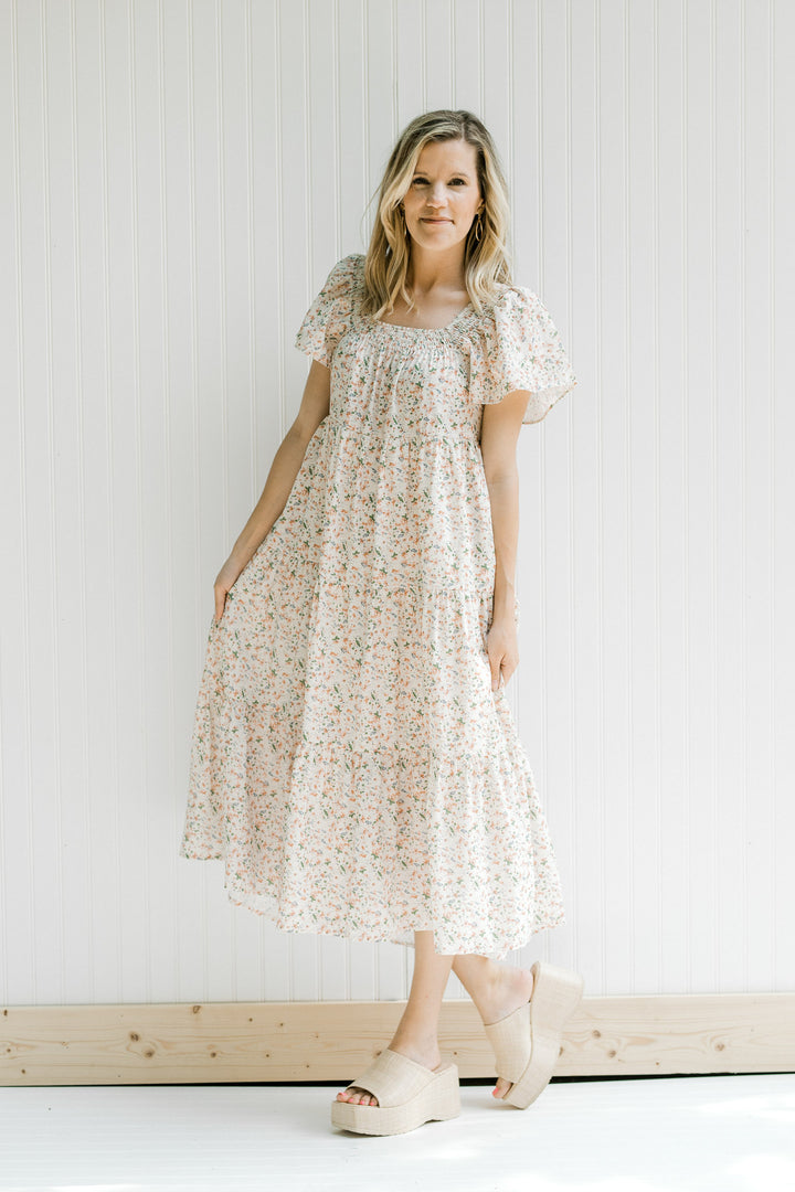 Blonde model wearing a tiered cream dress with a pink floral pattern and short flutter sleeves.