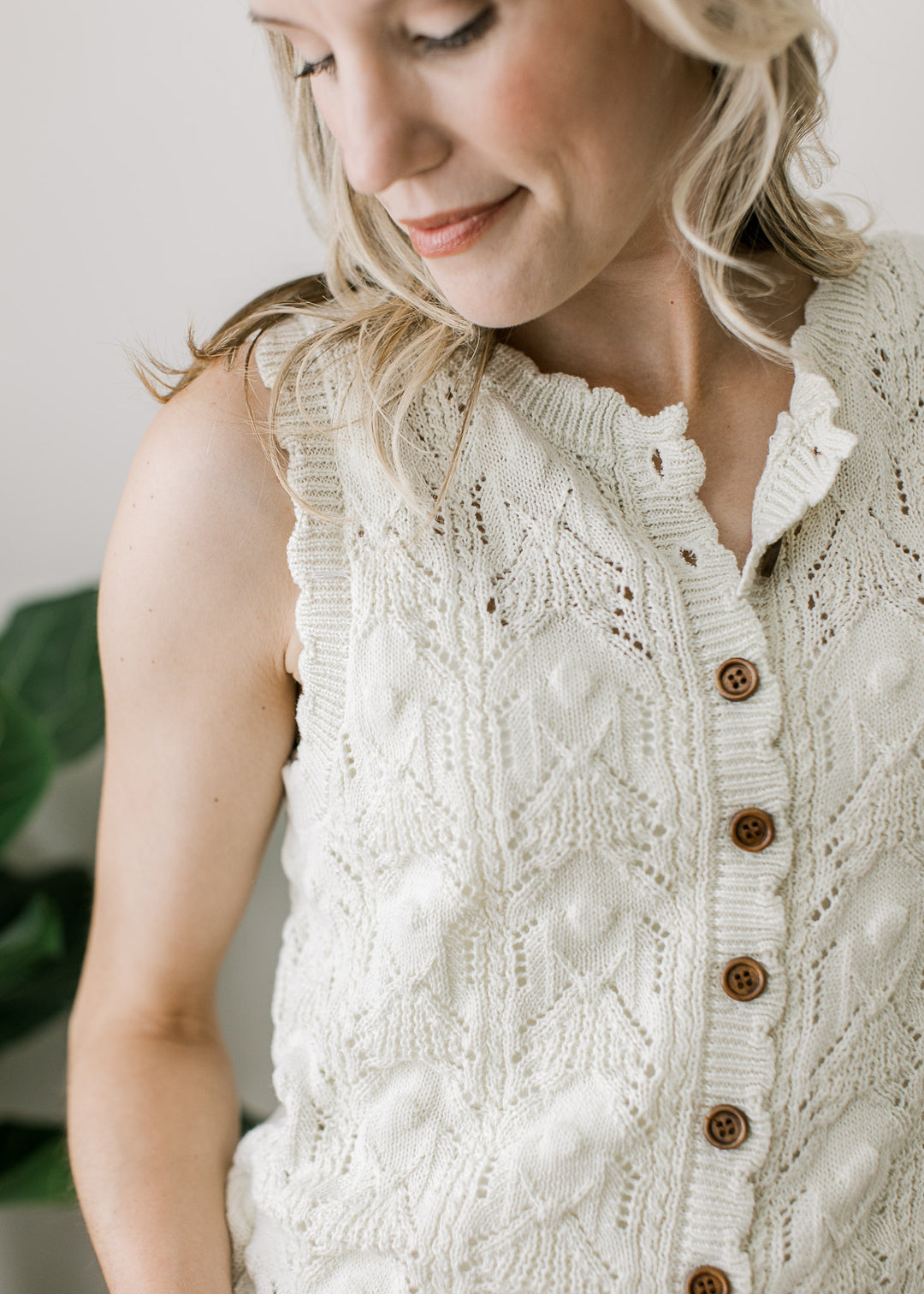 Model wearing a sleeveless cream knit sweater with brown buttons, scalloped neck, arm, and hem. 