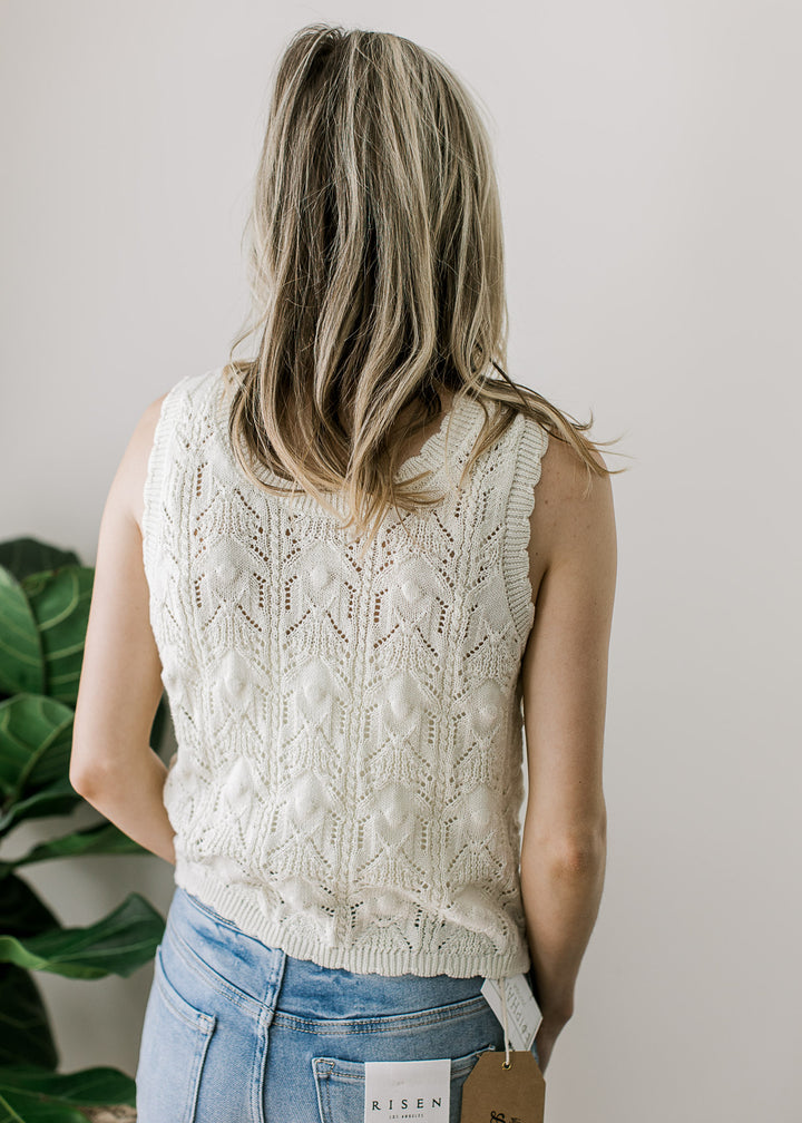 Back view of Model wearing a cream knit sweater with brown buttons, scalloped neck, arm, and hem. 