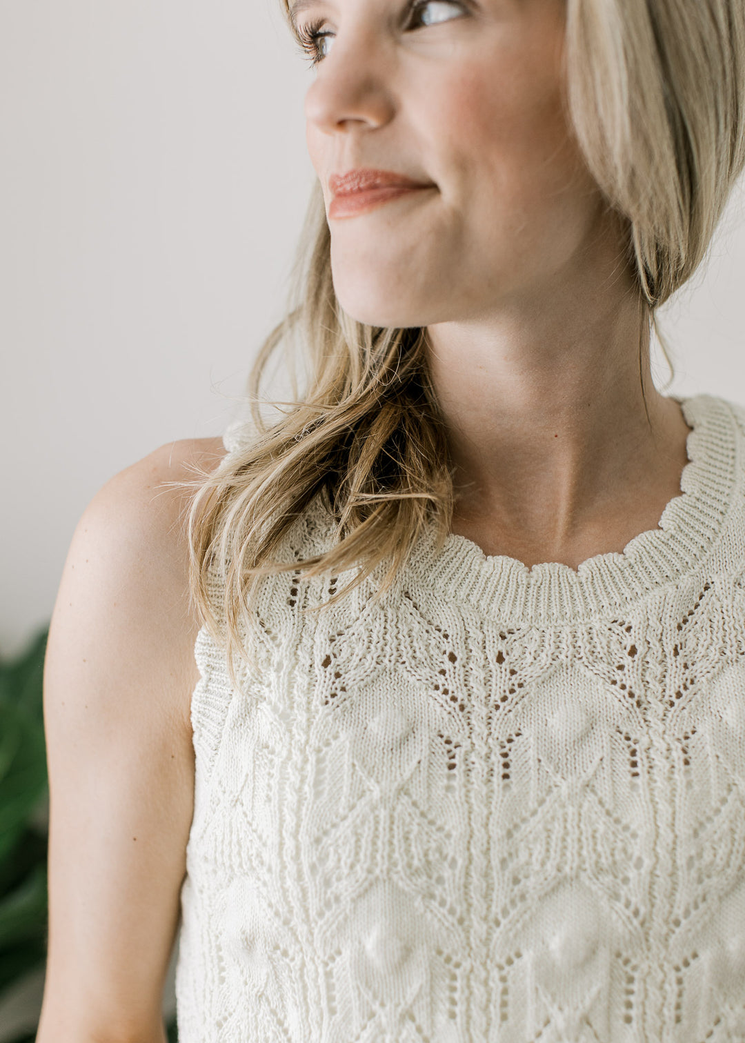 Close up scalloping at neck and arm of a cream knit sleeveless sweater with brown buttons. 